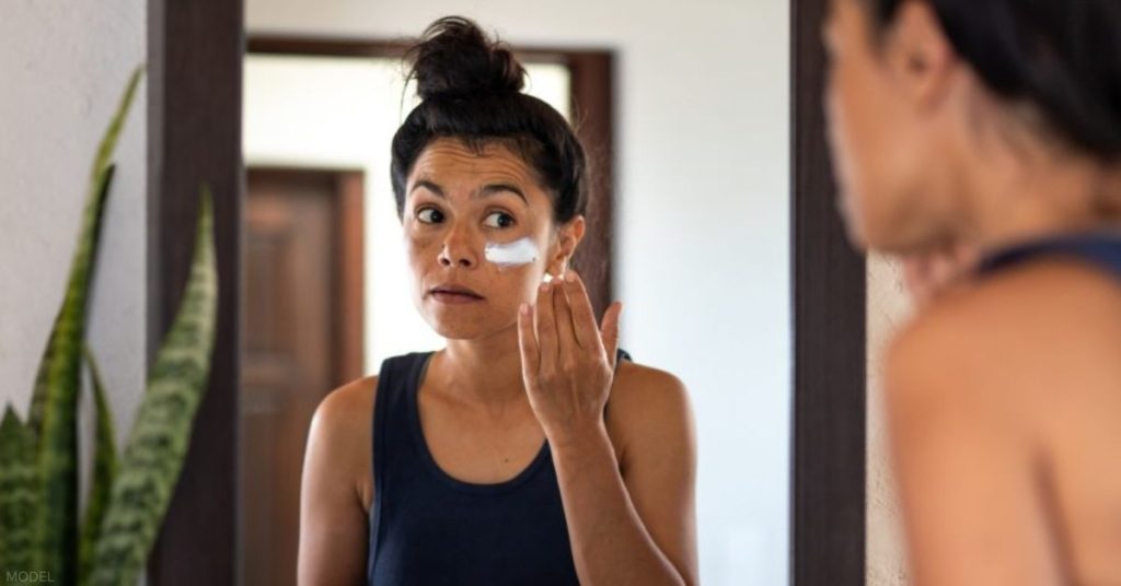 Woman applying white sunblock to face, looking at bathroom mirror (model)